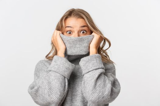 Close-up of cute blond girl, looking surprised, pulling grey sweater on her face, standing against white background.