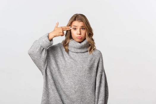 Portrait of sad blond girl in grey sweater, making finger gun sign near head as blowing her head, standing against white background.