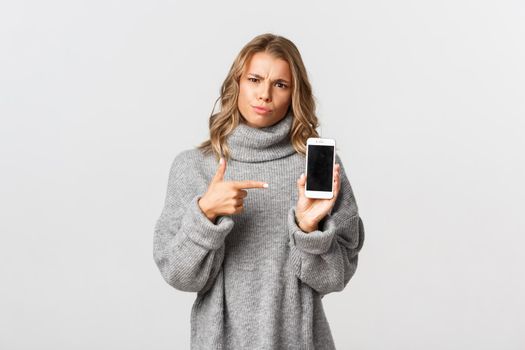 Portrait of angry girl pointing finger at smartphone, showing something disappointing and waiting for explanation, standing over white background.