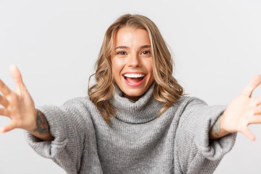 Close-up of happy attractive blond girl smiling, stretching hands forward for hug, reaching for something, standing over white background.