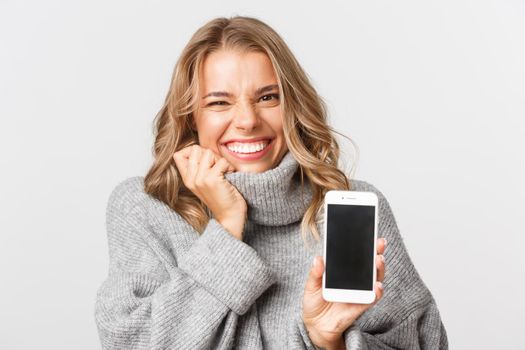 Close-up of happy and excited blond girl, smiling while showing mobile phone screen, standing over white background.