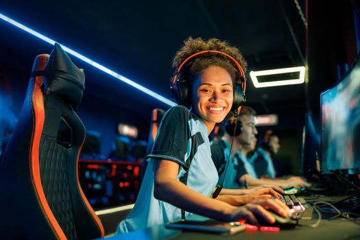African female gamer wearing headset looking at camera and smile while sitting in front of PC monitor in gaming room