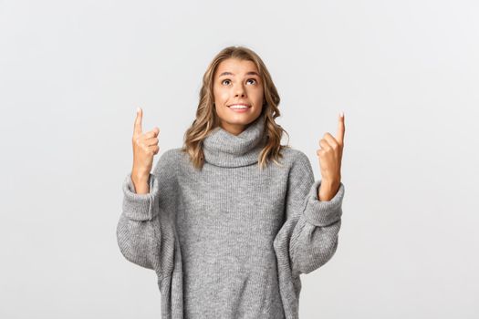 Image of hopeful young woman in grey sweater, pointing and looking up with happy smile, standing over white background.