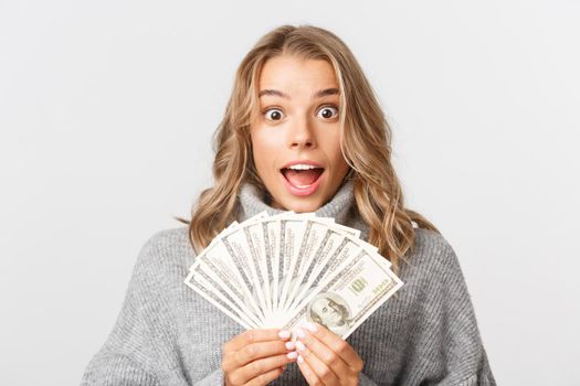 Close-up of excited rich girl with blond short hair, holding money and smiling amazed, standing over white background.