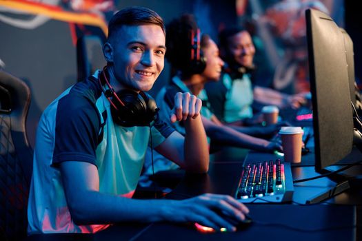Young man looking at camera and smiling while sitting in gaming chair in interior of dark cyber sports club