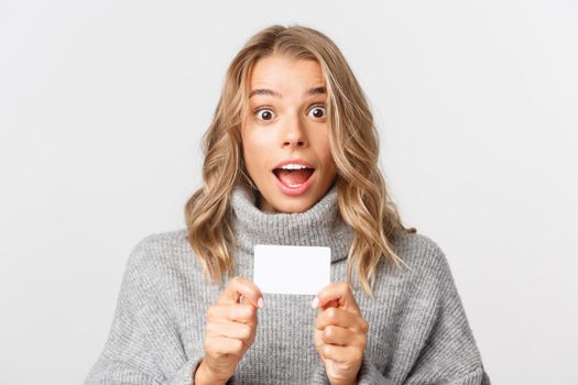 Close-up of attractive female model in grey sweater, looking amazed and showing credit card, standing over white background.