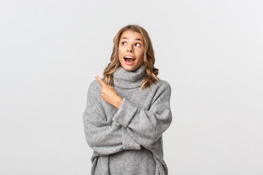 Portrait of amazed blond girl in grey sweater, open mouth and looking fascinated at your logo, pointing finger at upper left corner, white background.