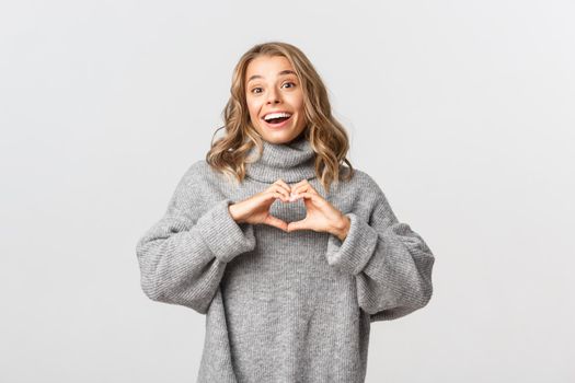 Image of romantic beautiful woman in grey sweater, looking passionate and showing heart sign, like something, standing over white background.