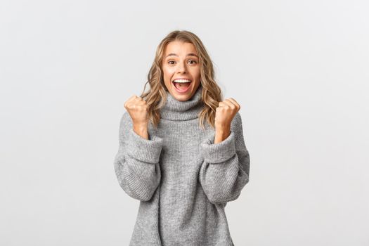 Image of successful happy woman saying yes, making fist pump and rejoicing from victory, triumphing over achievement, standing against white background.
