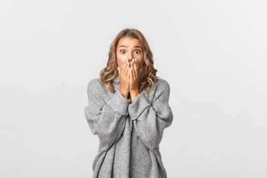 Close-up of shocked blond girl in grey sweater, gasping and looking concerned, standing over white background.