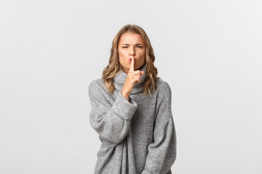 Close-up of angry blond girl shushing at someone, telling to be quiet, standing in grey sweater over white background.