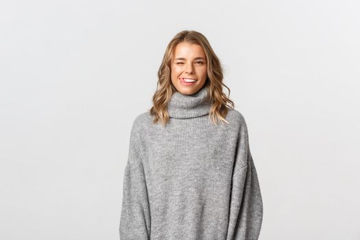 Sassy attractive blond girl in grey sweater winking, showing tongue and flirting with someone, standing over white background.