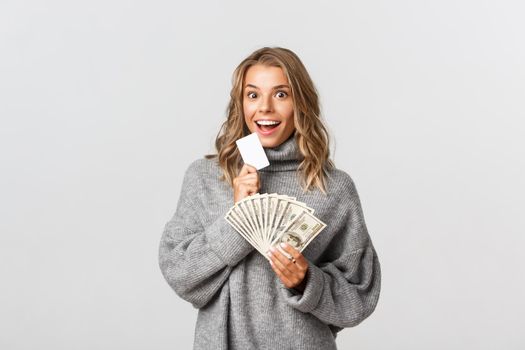 Studio shot of excited attractive girl holding money and credit card, smiling at camera, standing over white background.