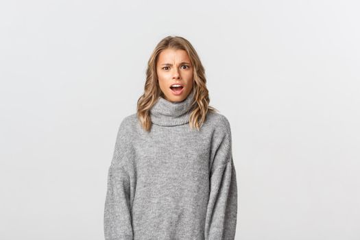 Close-up of confused blond girl in grey sweater, open mouth and looking puzzled at camera, standing over white background.