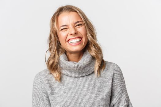 Close-up of happy beautiful girl with blond hair, wearing grey sweater, smiling and laughing, standing over white background.