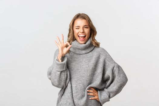 Confident smiling woman showing no problem gesture, make okay sign and winking self-assured, standing over white background.