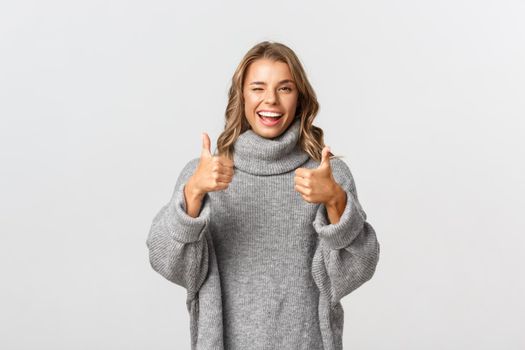 Image of attractive confident woman in grey sweater, showing thumbs-up to praise good choice, looking satisfied and winking, standing over white background.