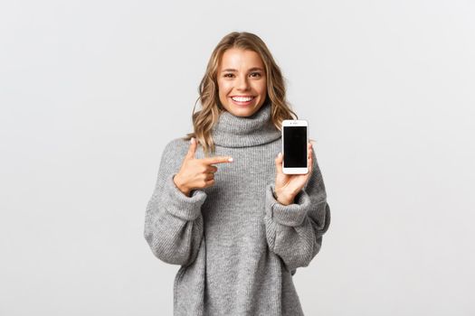 Portrait of attractive blond girl in grey sweater, pointing finger at mobile phone screen and smiling, recommending something.