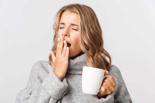 Close-up of sleepy blond girl in grey sweater, drinking coffee and yawning, standing over white background.