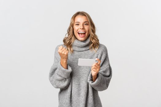 Image of happy winning girl in grey sweater, saying yes and triumphing, holding mobile phone, achieve goal in smartphone application, standing over white background.