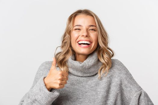 Close-up of happy blond girl in grey sweater, showing thumbs-up and smiling, like something, standing over white background.