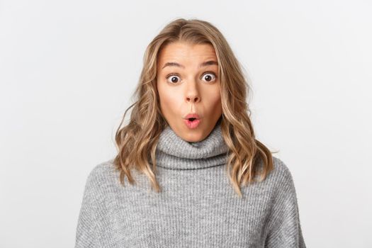 Close-up of attractive blond girl looking surprised, gasping amazed at camera, standing over white background.