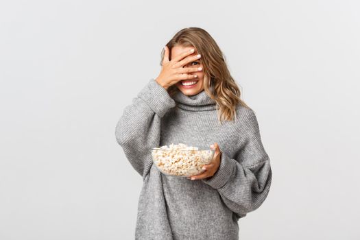 Happy cute girl with blond short hairstyle, watching movie and eating popcorn, looking through fingers at something embarrassing, standing over white background.
