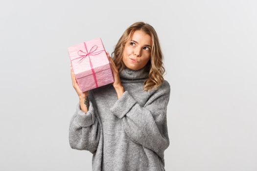 Image of cute blond girl shaking box with a gift, trying to guess what inside, standing over white background.