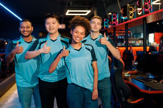 Happy multiracial cybersport team smiling at camera and showing thumbs up after esports tournament in gaming club