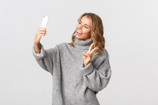 Portrait of attractive young blond girl in grey sweater holding smartphone, showing peace sign and taking selfie, standing over white background.
