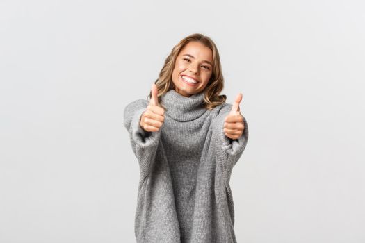 Image of happy beautiful woman in grey sweater, showing thumbs-up and smiling, give approval or agree, like and praise good choice, standing over white background.