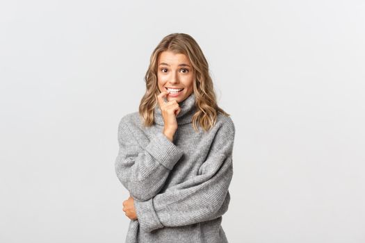 Image of nervous girl in grey sweater, biting finger and looking indecisive, standing over white background.