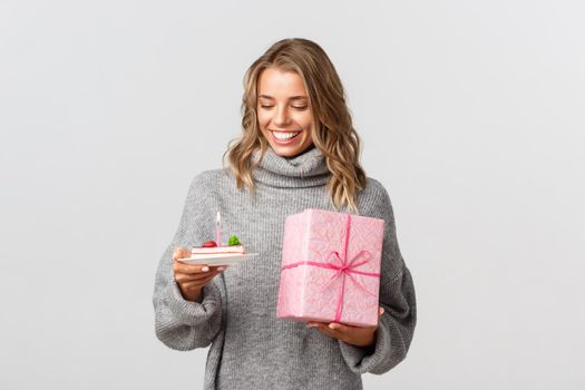 Studio shot of happy blond female model in grey sweater, looking at birthday cake, holding gift, celebrating b-day, standing over white background.