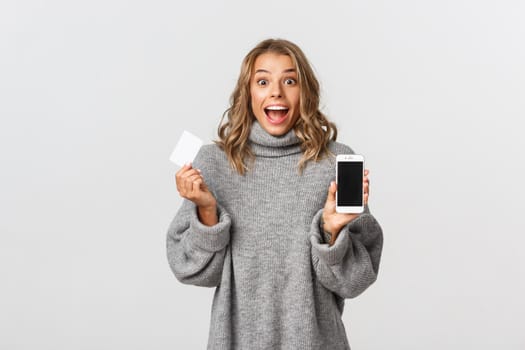 Excited beautiful girl with blond short hairstyle, showing smartphone screen and holding credit card, standing over white background.