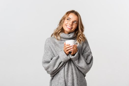 Image of dreamy smiling girl, looking at upper left corner thoughtful and drinking coffee, standing over white background.