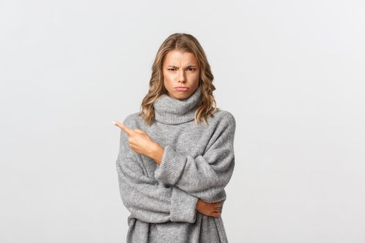 Image of disappointed moody girl, wearing grey sweater, sulking and frowning upset, pointing finger at upper left corner, standing over white background.