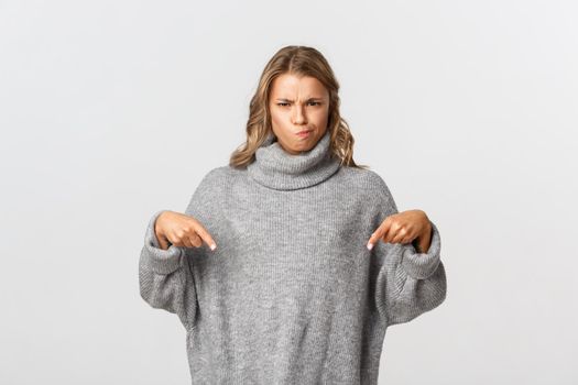 Image of doubtful and skeptical blond girl, frowning and pouting displeased, pointing fingers down at something bad, standing over white background.