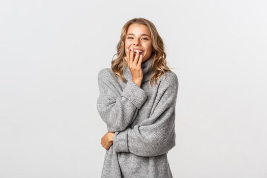 Image of attractive blond girl in casual clothing, laughing and cover mouth coquettish, standing against white background.