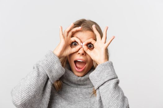 Close-up of happy attractive girl having fun, looking through finger glasses and look amazed, standing over white background.
