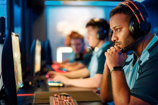 Side view of focused African professional gamer wearing headphones looking at PC screen while sitting in gaming club