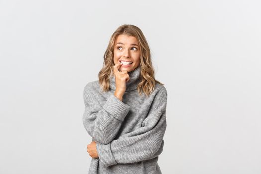 Image of indecisive nervous girl in grey sweater, biting finger and looking aside, standing over white background.