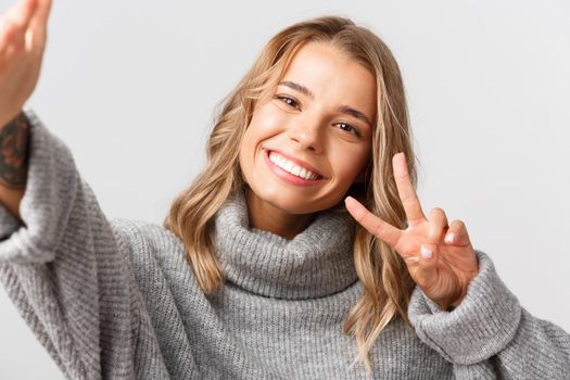 Close-up of attractive blond girl in grey sweater, taking selfie or having video call, showing peace gesture at mobile camera, standing over white background.