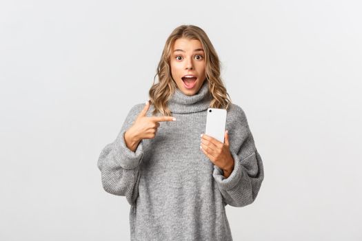 Portrait of amazed blond girl in grey sweater, pointing finger at mobile phone, recommend look at something online, standing over white background.