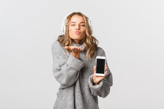 Young lovely woman in grey sweater, showing mobile phone screen, sending air kiss, standing over white background.