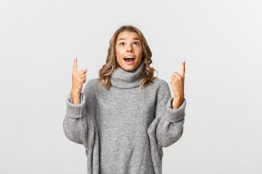 Image of surprised and happy blond girl, wearing grey sweater, pointing fingers up and looking fascinated, white background.