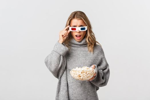 Image of blond girl in 3d glasses, holding bowl with popcorn, watching movie and looking confused, standing over white background.