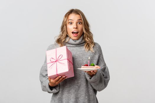 Portrait of attractive blond girl celebrating her birthday, holding gift and b-day cake, standing over white background.