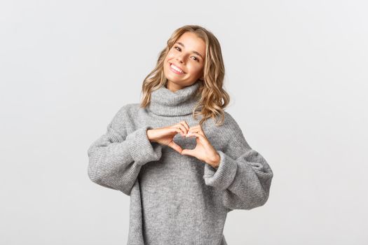 Image of lovely blond woman in grey sweater, smiling and showing heart sign, like something, standing over white background.