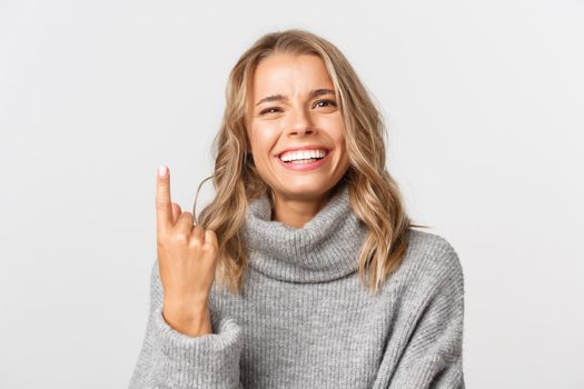 Close-up of carefree blond girl in grey sweater, showing one finger and smiling, standing over white background.
