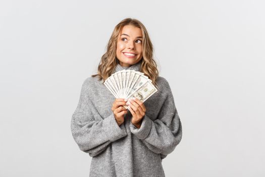 Portrait of dreamy cute girl in grey sweater, looking at upper left corner thoughtful and smiling, holding money, white background.
